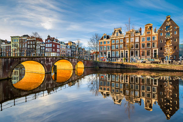 Sonniger Morgen an einer Gracht in Amsterdam, Niederlande, mit der Sonne scheinend unter einer Bogenbrücke von Michael Abid